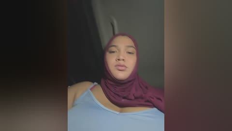 Media: Video of a young woman with light skin, wearing a light blue tank top and a maroon hijab, her head tilted slightly to the left, in a dimly lit room.