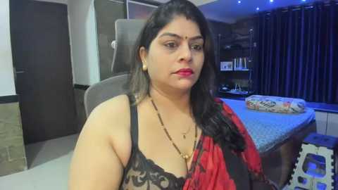 Media: Video of a plus-sized South Asian woman with long black hair, wearing a black lace bra and red sari, sitting on a chair in a modern, dimly-lit bedroom with a bed and dark curtains.