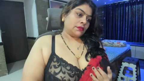 Media: Video of a South Asian woman with medium skin tone, long black hair, and red lipstick, wearing a black lace bra, holding a red bra, in a modern bedroom with dark curtains and a bed.