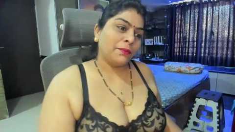 Media: Video of a South Asian woman with medium skin tone, dark hair, wearing a black lace bra, sitting on a bed in a modern bedroom with wooden furniture, curtains, and decorative pillows.