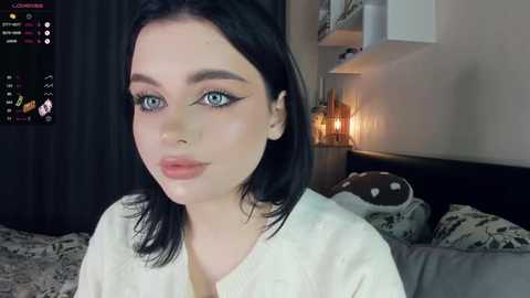 Media: Video of a young woman with pale skin, dark hair, and blue eyes, wearing white clothing, sitting on a bed in a dimly lit room.