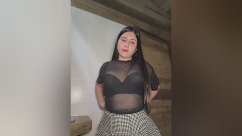 Media: Video of a young Latina woman with long black hair, wearing a sheer black top and checkered skirt, standing indoors near a brick wall.