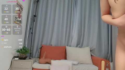 Media: Video of a nude woman with light skin, standing in a modern living room with grey curtains, orange couch, and a green potted plant.