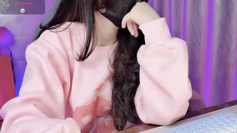 Media: Video of a person with long, wavy black hair, wearing a pastel pink sweater, partially obscuring their face with a black mask. Background features a purple-lit curtain and a computer keyboard.