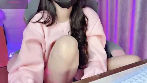 Media: Video of a woman with long black hair, wearing a pink sweater, black mask, and seated at a computer, with a purple-lit background.