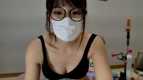 Media: Video of an East Asian woman with glasses and a white mask, wearing a black bra, sitting in a colorful room with a sewing machine and fabric.