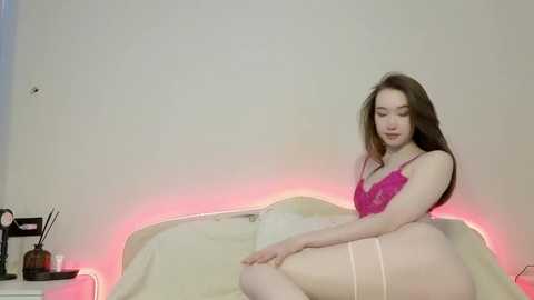 Media: A video of a young, fair-skinned woman with long brown hair, wearing a pink lace bra and matching thong, sitting on a bed with a cream-colored blanket, illuminated by soft pink LED lights.