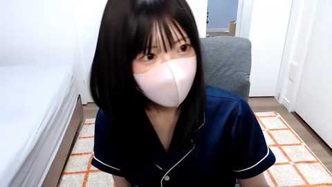 Media: A video of an Asian woman with shoulder-length black hair, wearing a blue shirt and a white face mask, seated on a beige and orange-patterned rug in a minimalist bedroom with white walls and a bed.