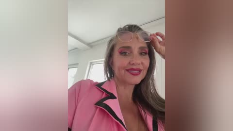 Media: Video of a fair-skinned woman with long brown hair, wearing pink lipstick, pink eyeshadow, and a pink jacket with black trim, adjusting her glasses in a bright, modern room with white walls and windows.