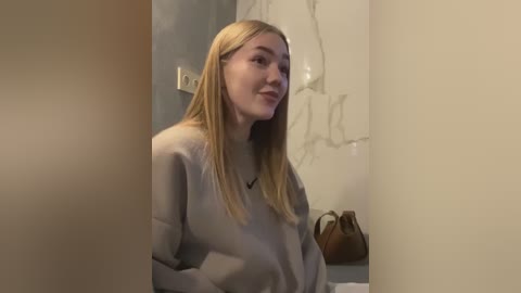 Media: Video of a young woman with long blonde hair, wearing a light grey sweater, standing in a dimly lit room with beige walls and a brown handbag.