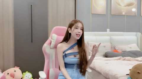 Media: Video of an Asian woman with long brown hair, wearing denim overalls, seated on a pink gaming chair in a modern bedroom with white bedding and floral artwork.