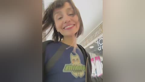 Media: Video of a young woman with short brown hair, smiling, wearing a Batman shirt, standing in a brightly lit, colorful room with a TV in the background.