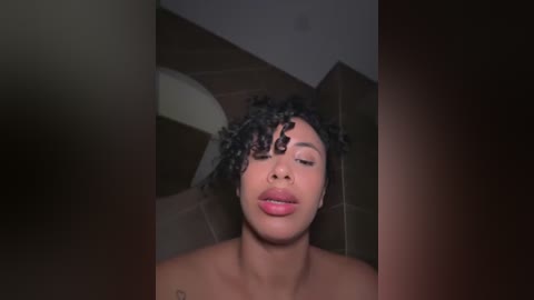 Media: Video of a young Black woman with curly hair, closed eyes, and full lips, taken in a dimly lit bathroom with a tiled shower wall in the background.