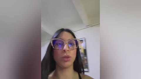 Media: Video of a young woman with long black hair and large, round glasses, standing indoors with a white wall and abstract artwork in the background.