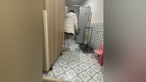 Media: Video of a woman in a white puffer jacket and knee-high socks walking away from a bathroom with white floral-patterned floor tiles and a pink chair.