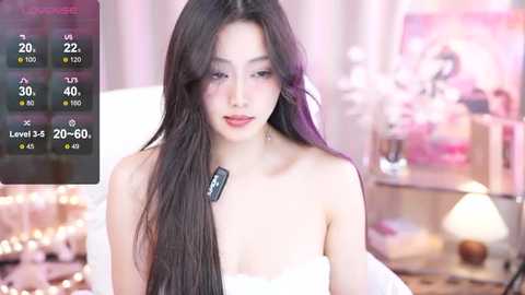 Media: Video of an Asian woman with long black hair, wearing a white strapless dress, sitting indoors with a blurred background.