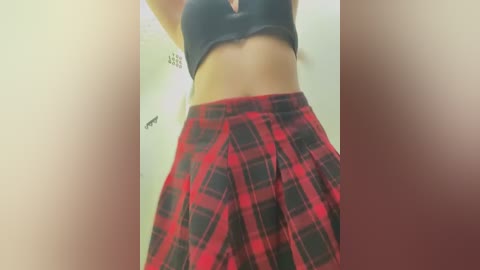 Media: Video of a woman's midsection in a black bra and high-waisted, red and black plaid skirt. She has a medium build with a flat stomach, standing in a bathroom with a tiled wall and a small, black-framed mirror.