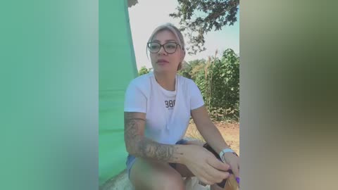 Media: Video of a young, fair-skinned woman with short, platinum blonde hair and glasses, wearing a white t-shirt and shorts, sitting outdoors near a green and white tent.