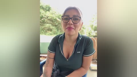 Media: Video of a middle-aged woman with light skin, glasses, and short hair, wearing a dark green polo shirt, sitting outdoors with a blurred background of greenery.