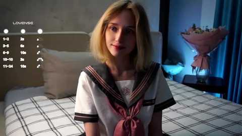 Media: Video of a young blonde woman in a school uniform, standing on a bed with a checkered white and black bedspread in a dimly lit bedroom.