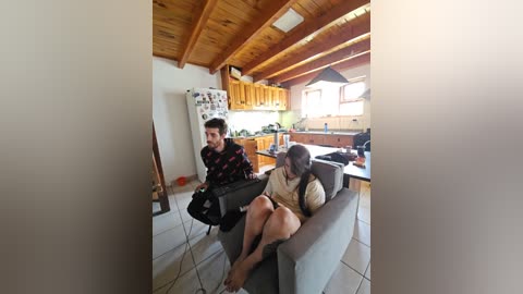 Media: Video of a cozy kitchen with wooden ceiling and cabinets, featuring a man and woman seated on a gray couch, engaging in conversation.