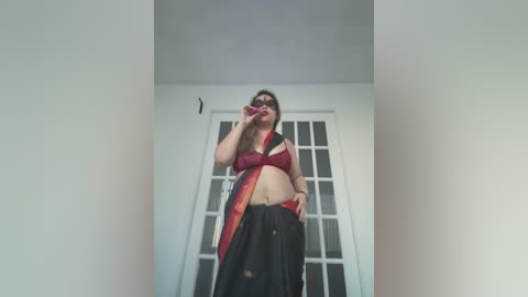 Media: Video of a curvy woman with light skin, wearing a red lace bra, black skirt, and sunglasses, posing seductively in a dimly lit room with white walls and glass doors.