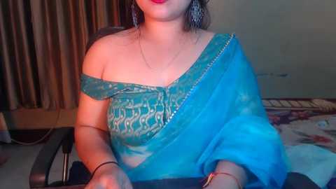 Media: Video of a South Asian woman with fair skin, wearing an off-shoulder blue saree with gold embroidery, seated on a chair.