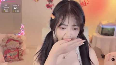 Media: Video of a young Asian woman with long black hair and pigtails, wearing a white tank top, holding a phone, in a cozy room with decorations and a fish tank.