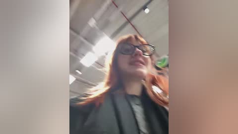 Media: A video captures a woman with long, reddish-brown hair and glasses, wearing a dark jacket, standing in a brightly lit, industrial-looking room with white walls and exposed pipes.