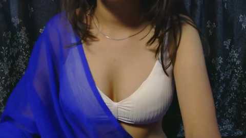 Media: Video of a light-skinned woman with long brown hair, wearing a white bra and blue sheer fabric draped over one shoulder, against a dark floral-patterned background.