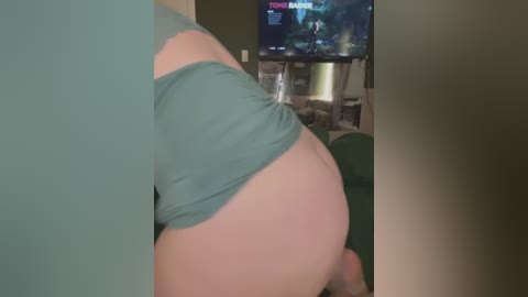 Media: Video of a person with fair skin and a large, bare buttocks in teal shorts, captured from a side angle. Background shows a living room with a green couch and a television displaying a video game scene.