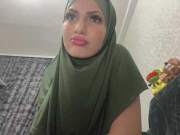 Media: Video of a light-skinned woman with a round face, wearing a green hijab and pink lipstick, standing in a room with a patterned wallpaper, a yellow toy, and a child playing nearby.