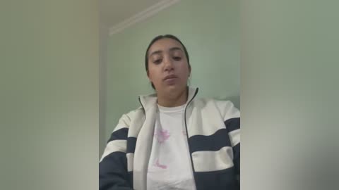 Media: Video of a young woman with medium-dark skin, brown eyes, and dark hair tied back, wearing a striped jacket and a white T-shirt with pink floral designs. She stands against a pale green wall in a simple room.