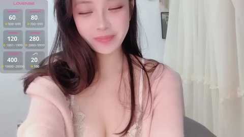 Media: Video of a young East Asian woman with long, straight black hair and fair skin, wearing a light pink cardigan over a white lace bralette, looking serene with her eyes closed. Background features a white wall and a partially visible window.