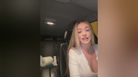 Media: Video of a blonde woman with long hair, wearing a white top, sitting in a car with a roll of toilet paper visible on the seat behind her.