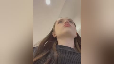 Media: Video of a young woman with fair skin and long brown hair, wearing a dark sweater, looking up from a low angle. The background is blurred, with a white ceiling light visible.