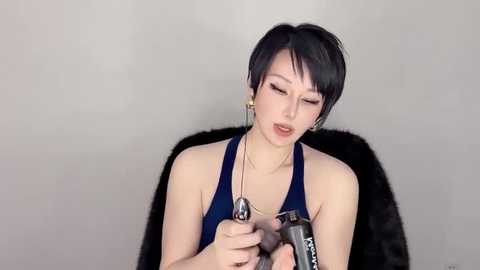 Media: A video of a young Asian woman with short black hair, wearing a blue halter top, holding a silver gun. She has a slender physique and a neutral expression. The background is plain and light-colored.