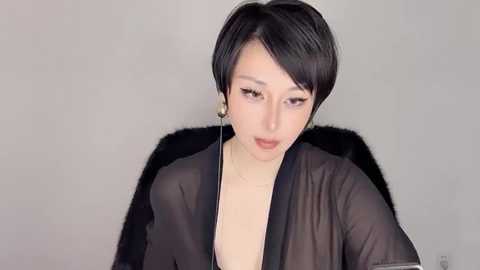 Media: Video of a young Asian woman with short, black hair, wearing a sheer black robe, sitting against a plain, light gray background. She has a fair complexion and wears long, dangling earrings.