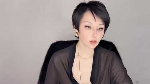 Media: Video of an East Asian woman with short black hair, wearing a sheer black robe, gold earrings, and a necklace, seated against a plain grey background.