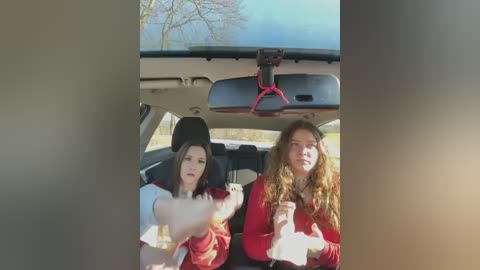 Media: Video of two women in a car, one with long curly hair, wearing a red sweater, the other with straight hair, wearing a red jacket, both looking out the window.