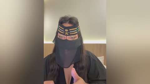 Media: A video of a woman with long dark hair, wearing a black sheer mask with intricate gold and white patterns, covering her eyes and nose, and a black robe. The background shows a beige wall and wooden furniture.