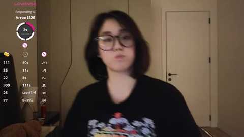 Media: A video of a young woman with glasses and shoulder-length black hair, wearing a black t-shirt with colorful graphics. She stands in a dimly lit room with beige walls and a closed door.