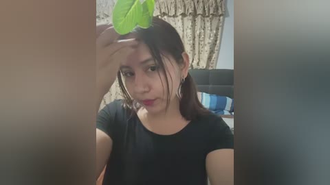 Media: Video of a young woman with light skin and dark hair, wearing a black shirt, holding a green leaf over her head in a playful pose. Background features a beige curtain and a blue and white patterned cushion.