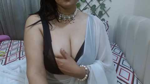 Media: Video of a woman with light skin, medium build, and dark hair, wearing a sheer white robe, black bra, and a necklace, holding her breast, in a bedroom with patterned bedding and a white couch.