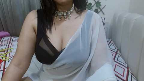 Media: Video of an Indian woman with fair skin, wearing a black bra and a semi-transparent white saree, adorned with a beaded necklace, seated on a bed with a geometric-patterned duvet.