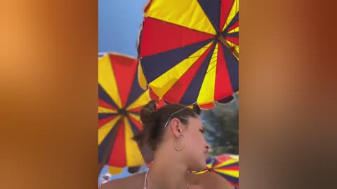 Media: Video of a young woman with brown hair in a bun, wearing a bikini and large hoop earrings, holding a large, colorful umbrella with a red, yellow, and blue pattern.