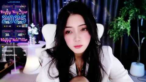 Media: Video of an Asian woman with long black hair, wearing a white jacket, seated indoors. Background features a potted plant and festive banners.