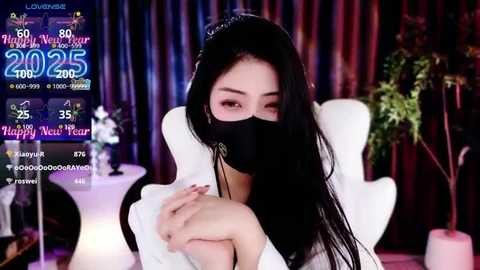 Media: Video of an Asian woman with long black hair, wearing a white dress, black mask, and a sparkling crown, sitting on a white chair in a dimly lit room with red curtains.