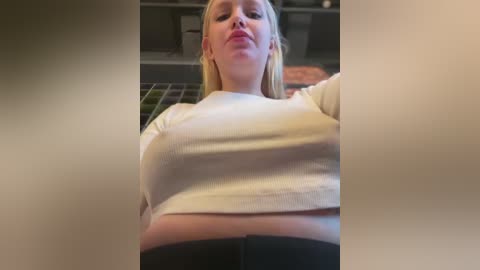 Media: A video of a blonde woman with fair skin and full lips, wearing a tight white top, taken from a low angle, showing her cleavage. The background is blurred, suggesting an indoor setting.