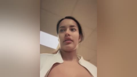 Media: Video of a woman with fair skin, dark hair, and a medium build, wearing a white blouse, looking slightly surprised in an office setting with beige walls and a fluorescent light.